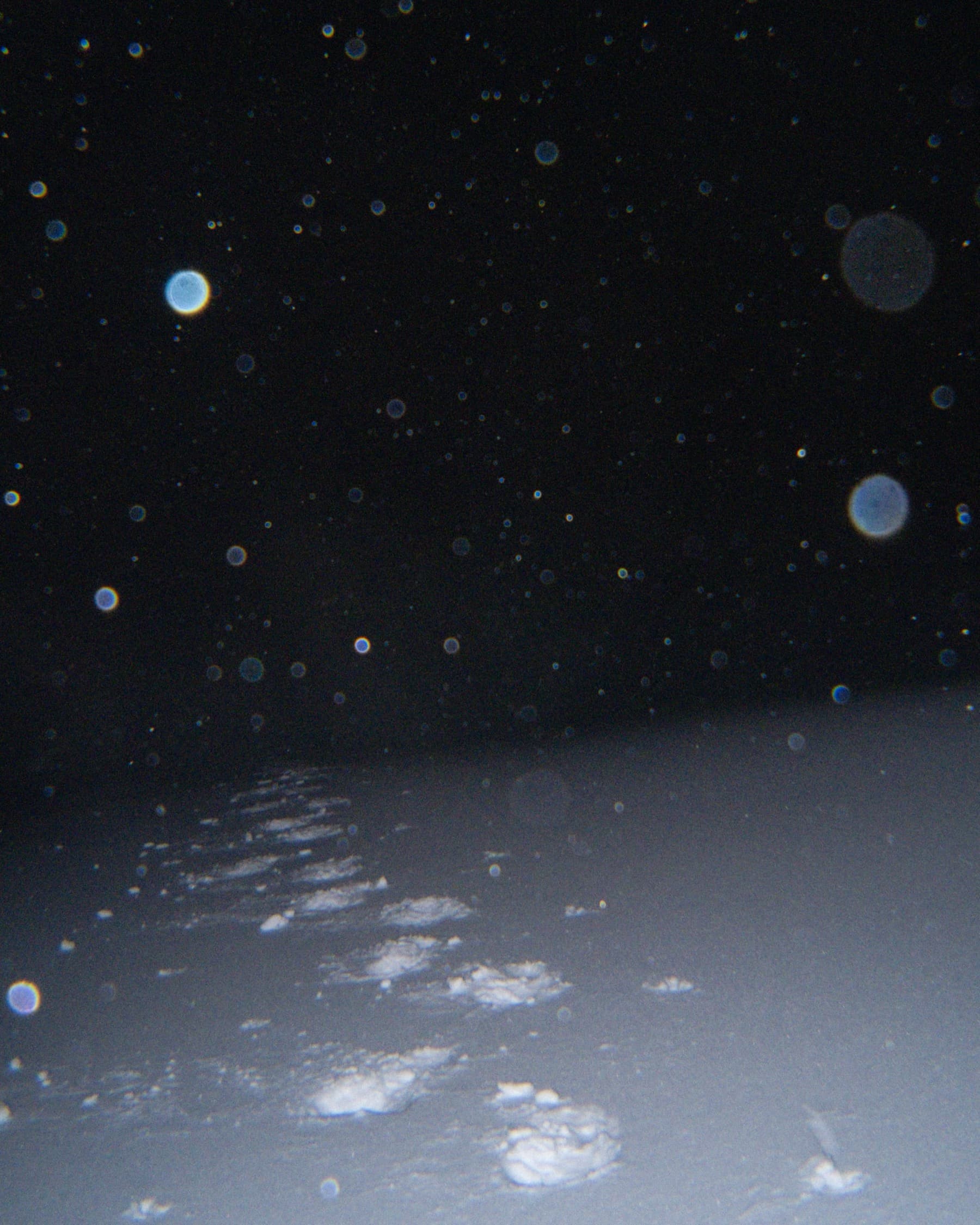 Snowflakes falling at night illuminated against a dark sky, with footprints in the snow visible at the bottom of the frame