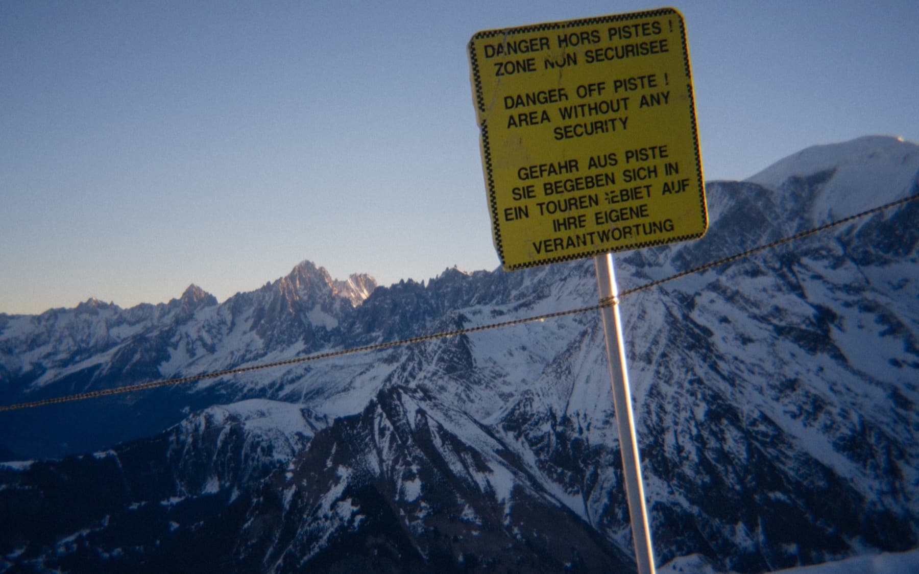 Off-piste danger warning sign in French, English, and German with dramatic snow-covered peaks and Mont Blanc massif visible behind at dusk