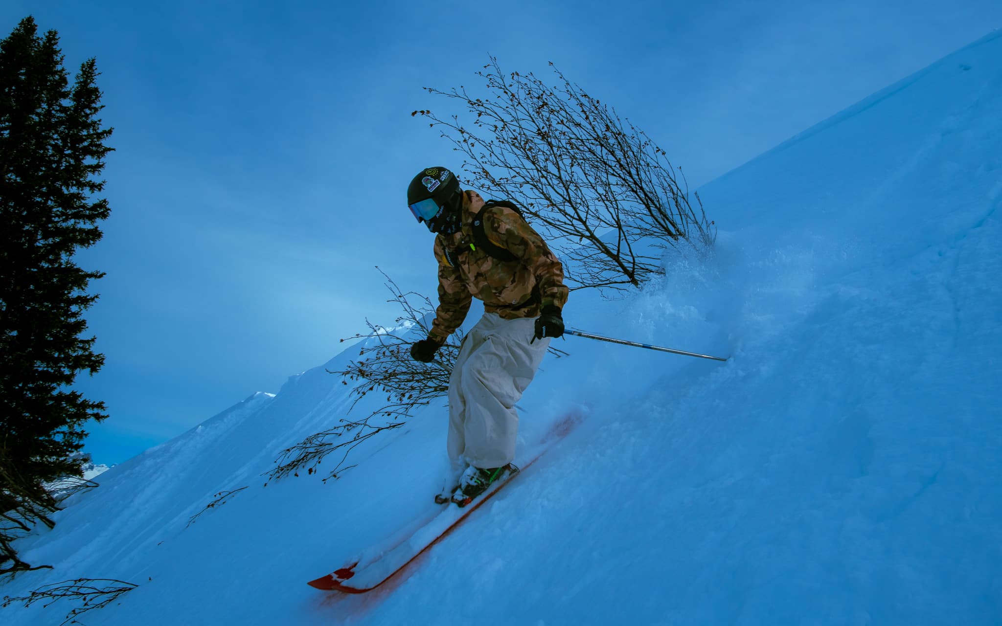 Skier in camo jacket carving through deep powder on a steep slope with pine trees and snow spray