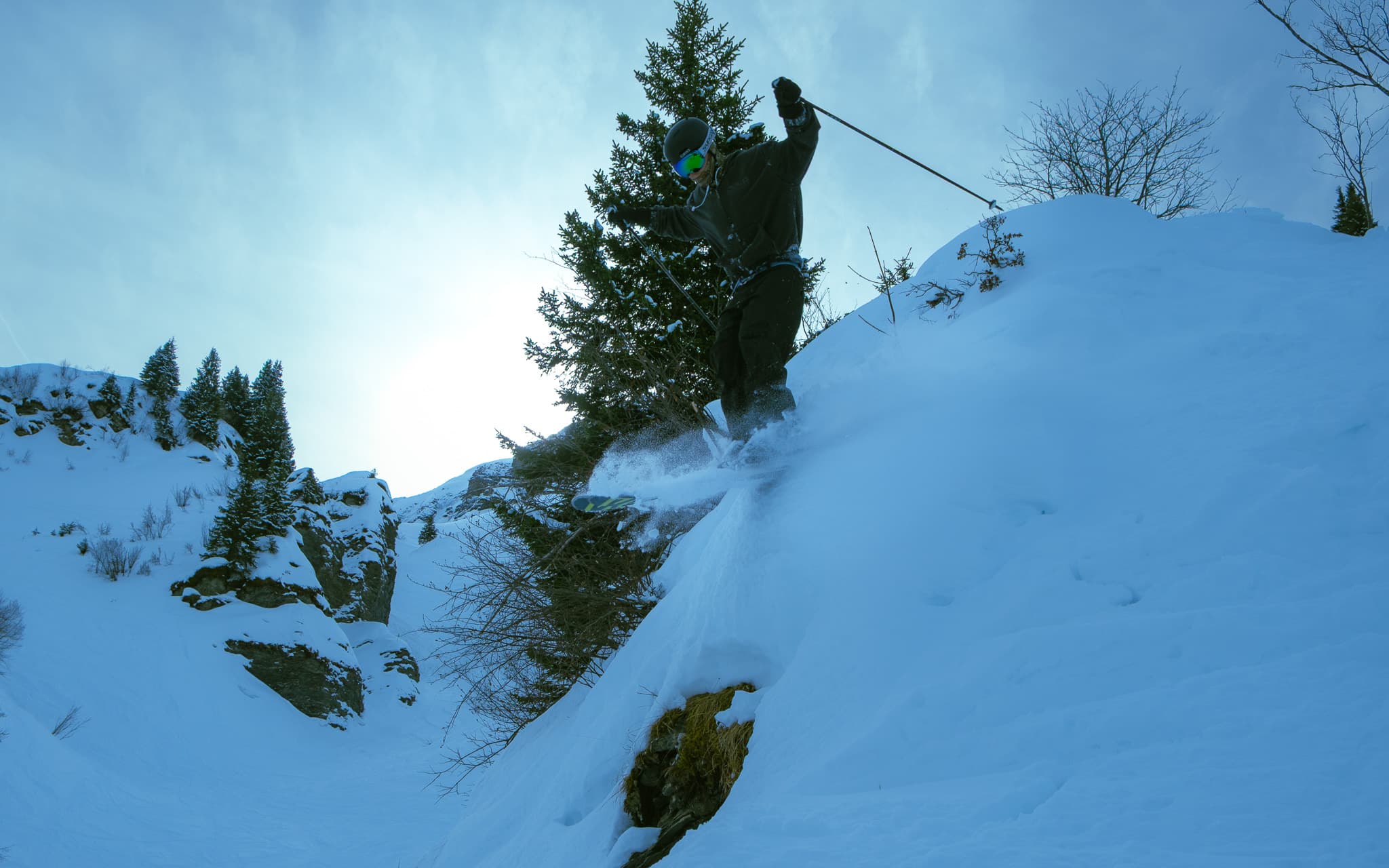 Skier dropping off a rocky cliff edge into powder snow with pine trees and mountains behind