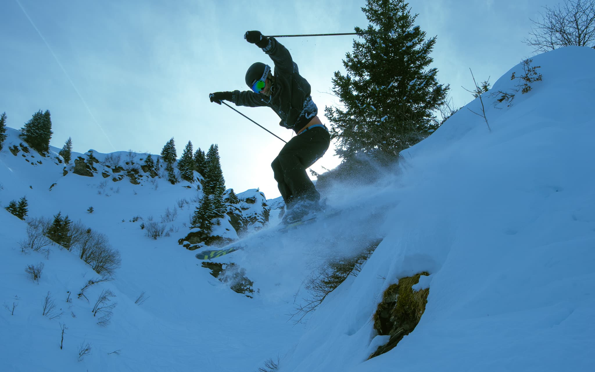 Skier jumping off a rock drop with poles raised and snow spraying beneath against a backdrop of snowy pines