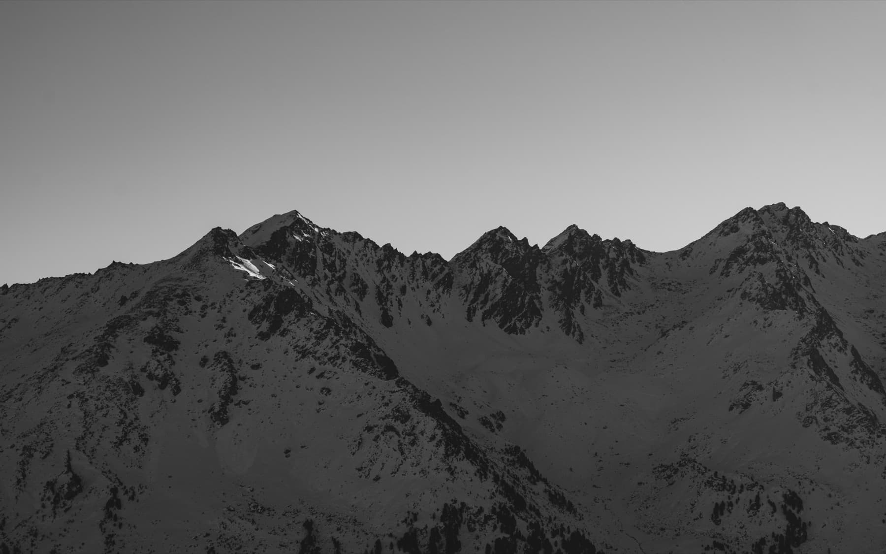 Jagged mountain ridge with multiple peaks silhouetted against a grey sky, minimal snow on the dark rocky faces, shot in black and white