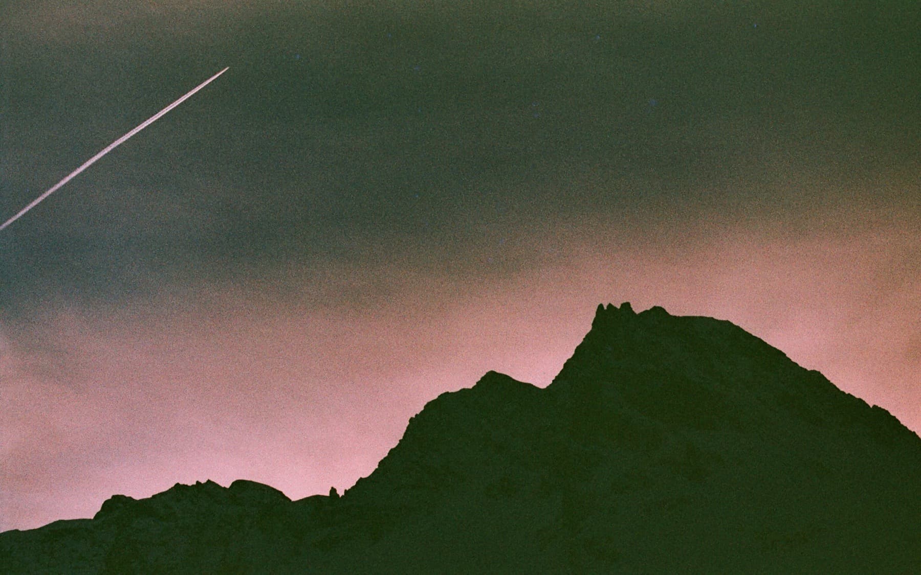 Silhouette of a jagged mountain ridge against a dusky pink and teal sky with a single airplane contrail streaking overhead