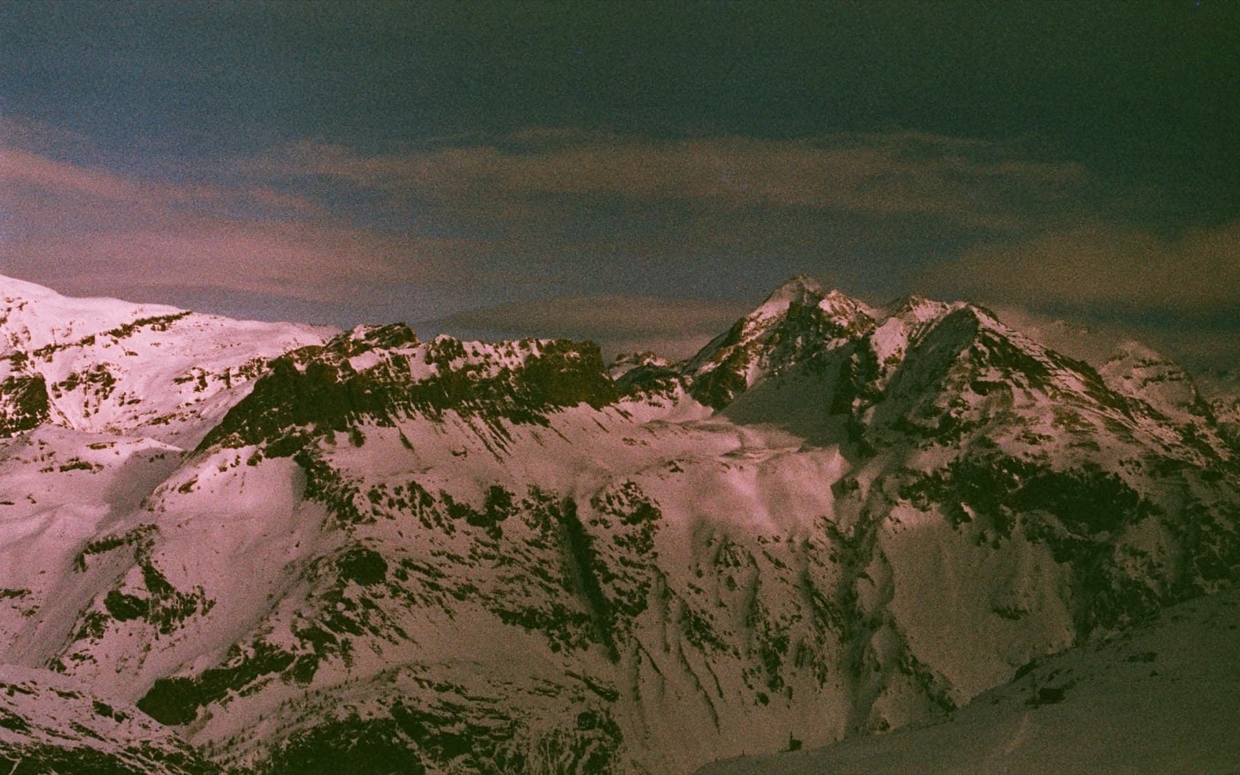 Panoramic view of rugged snow-covered high-altitude peaks with exposed rock faces under a moody pink-tinted sky