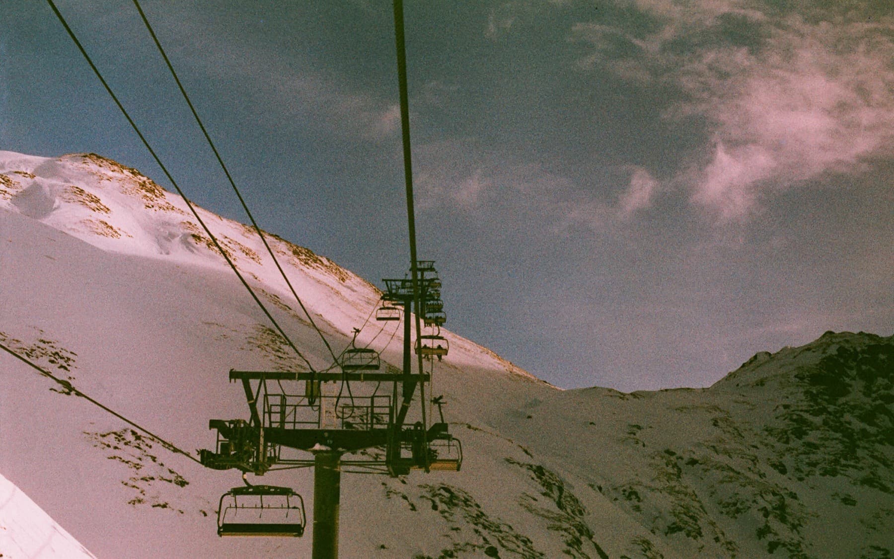 Empty chairlift ascending a steep snow-covered mountainside with cables stretching upward against a partly cloudy sky