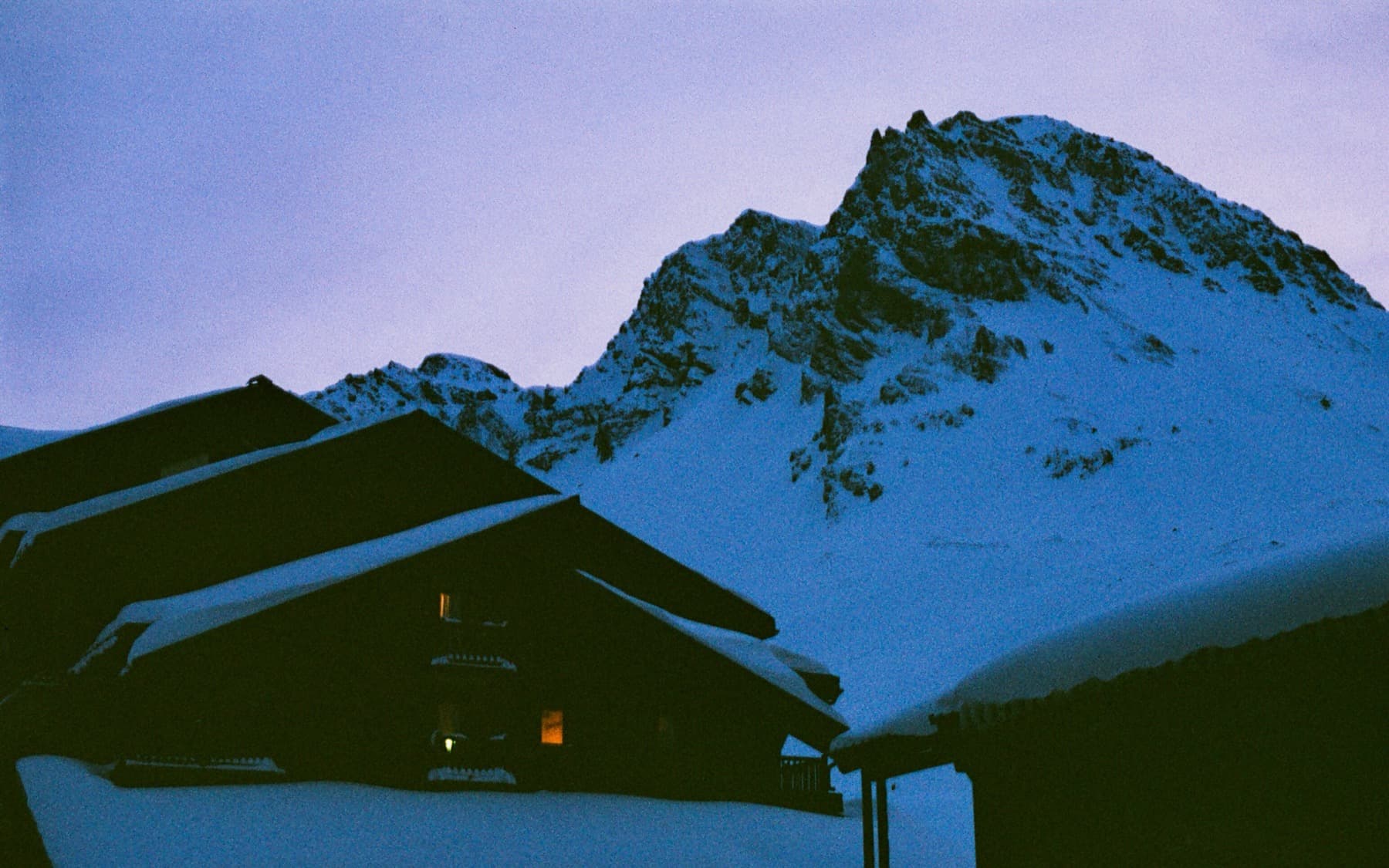 Snow-covered mountain chalet with glowing windows at twilight, dwarfed by a dramatic rocky peak rising steeply behind in blue evening light