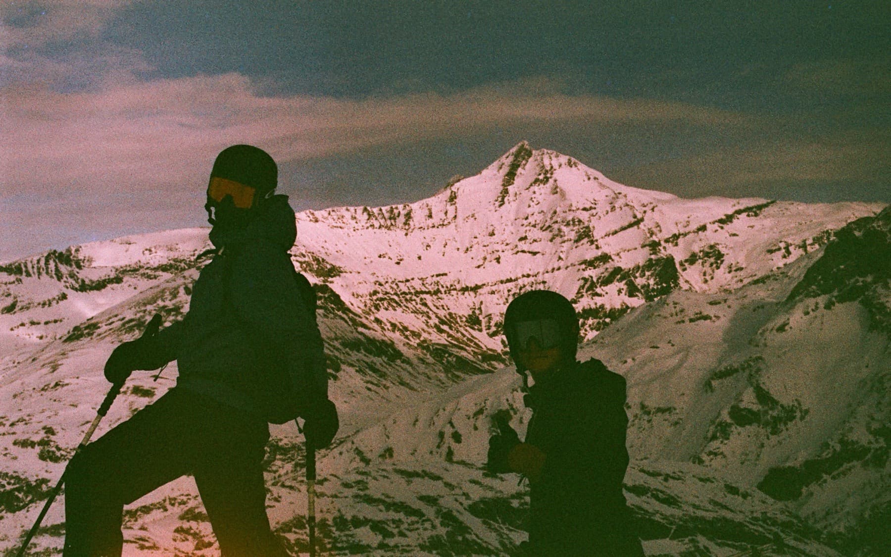 Two skiers silhouetted against a snow-covered peak bathed in pink alpine light, shot with grainy expired film tones