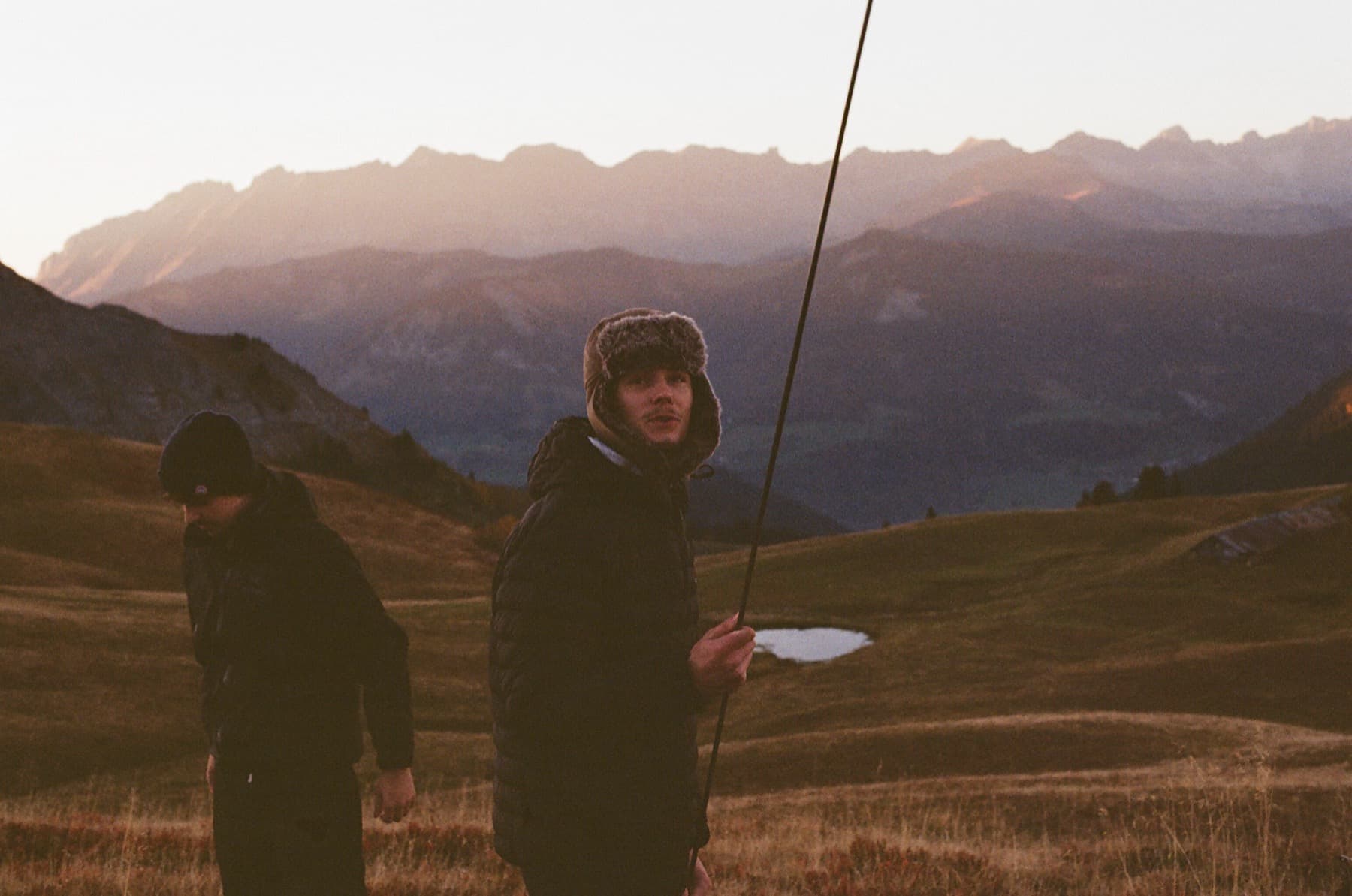 Two hikers in warm jackets on an alpine plateau at golden hour, one wearing a fur-lined hat, shot on film with warm tones and hazy mountains behind
