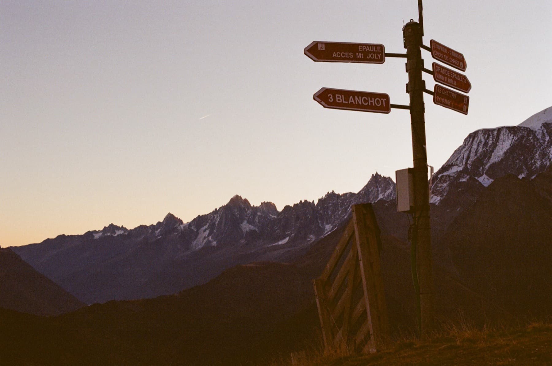 Trail signpost pointing to Mont Joly and Blanchot silhouetted against a dusky sky with jagged Alpine peaks and the Mont Blanc range on the horizon, shot on film