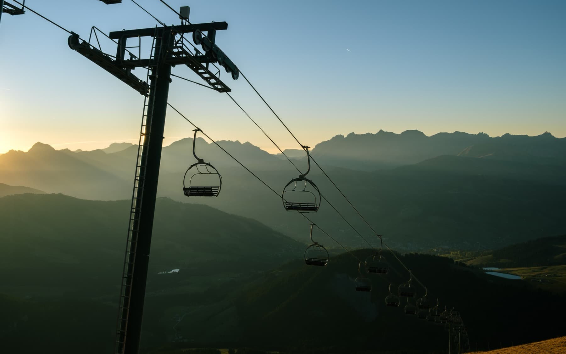 Silhouetted chairlift chairs hanging against a golden sunrise with hazy layered mountain ridges in the background