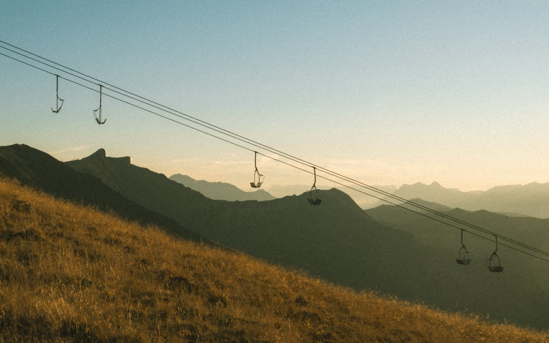 Empty ski lift chairs crossing a golden grassy hillside at sunrise with silhouetted mountain ridges receding into the haze