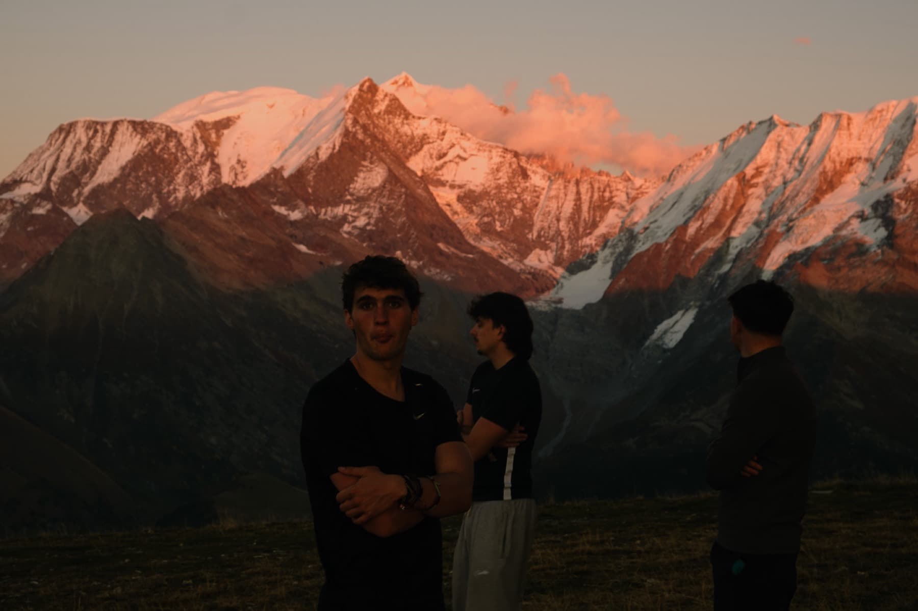 Three young men standing on an alpine ridge at dusk with the Mont Blanc massif glowing pink with alpenglow behind them